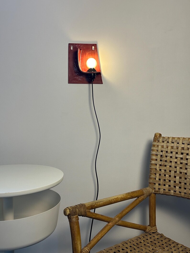 May include: A wall-mounted lamp featuring a rectangular, dark red ceramic backplate with a curved, bowl-like center. A black socket holds a glowing, round light bulb. A thin black power cord hangs vertically from the base of the light fixture.