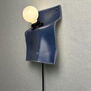 May include: A wall-mounted lamp featuring a curved, deep blue ceramic base with a glossy finish. A single round, frosted white light bulb is secured in a black socket attached to the ceramic. A thin black power cord hangs vertically from the base.