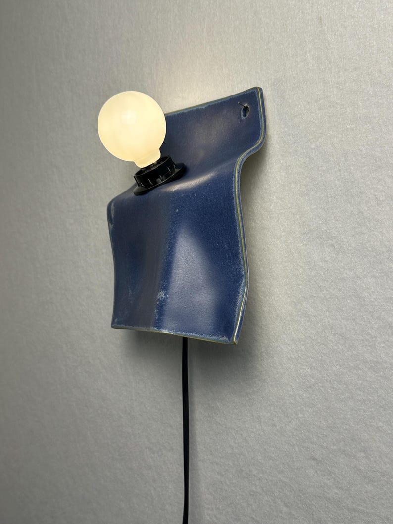 May include: A wall-mounted lamp featuring a curved, deep blue ceramic base with a glossy finish. A single round, frosted white light bulb is secured in a black socket attached to the ceramic. A thin black power cord hangs vertically from the base.