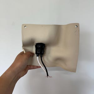 May include: A beige, rectangular wall-mounted light fixture base with a molded, undulating surface. It features a central black circular socket housing, two pre-drilled mounting holes at the top corners, and two exposed electrical wires.