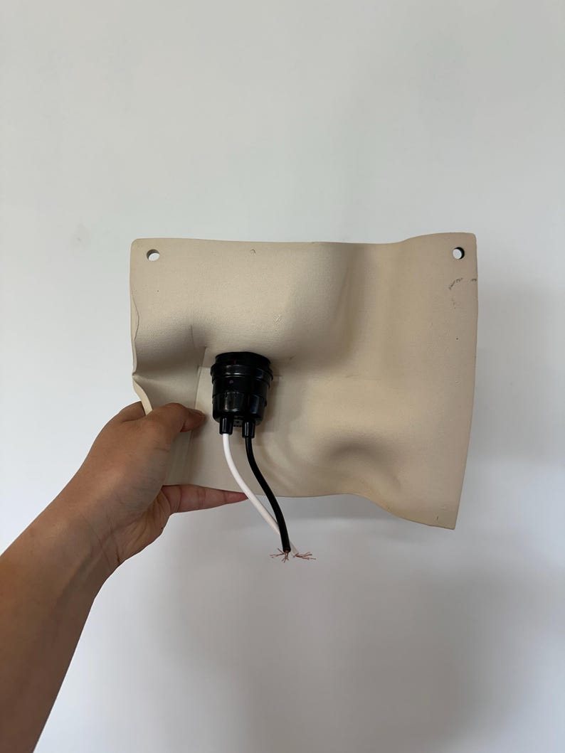 May include: A beige, rectangular wall-mounted light fixture base with a molded, undulating surface. It features a central black circular socket housing, two pre-drilled mounting holes at the top corners, and two exposed electrical wires.