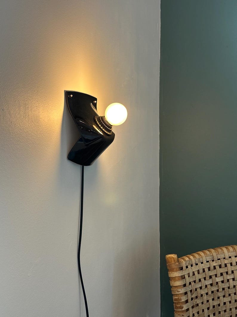 May include: A black wall-mounted lamp with a glossy finish and a sculptural, angular form. The fixture features a single round, glowing light bulb attached to a metallic socket, with a thin black power cord extending downward from the base.