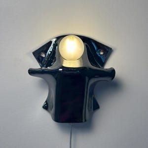 May include: A wall-mounted lamp featuring a glossy black ceramic base with a wide, flared shape and two mounting holes. A single, round, frosted glass light bulb is positioned at the top center, emitting a soft, warm glow against the wall.