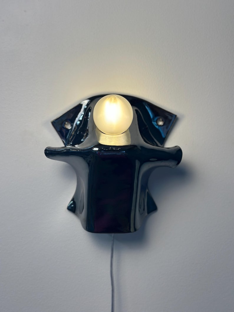 May include: A wall-mounted lamp featuring a glossy black ceramic base with a wide, flared shape and two mounting holes. A single, round, frosted glass light bulb is positioned at the top center, emitting a soft, warm glow against the wall.