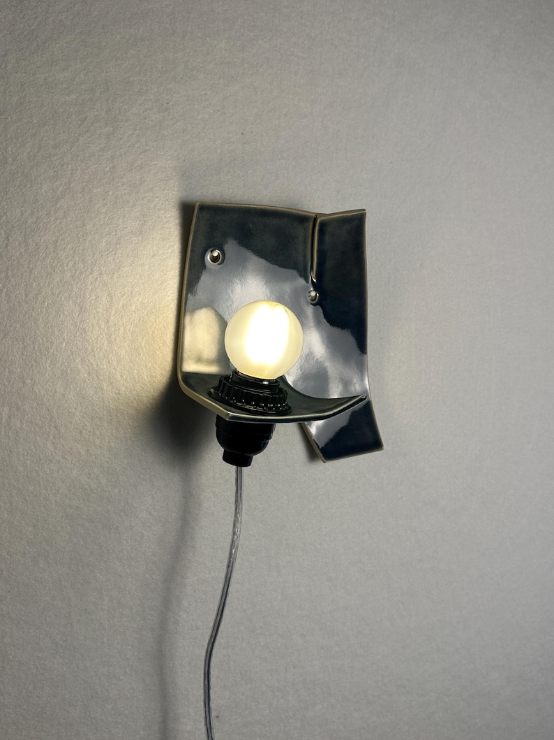 May include: A ceramic wall sconce with a dark blue glaze. The sconce has a unique, folded design with a small shelf to hold a round, glowing light bulb. A black socket and a gray cord complete the fixture.