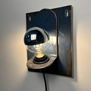May include: A wall-mounted lamp featuring a square, dark blue glazed ceramic backplate with a rounded, protruding shelf. A black socket holds a clear glass light bulb with a reflective silver-colored top half. A black power cord extends downward.