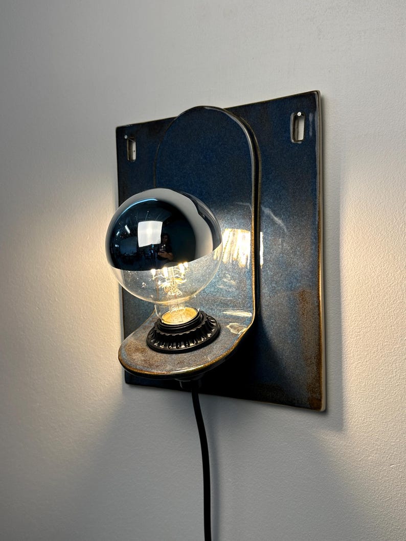 May include: A wall-mounted lamp featuring a square, dark blue glazed ceramic backplate with a rounded, protruding shelf. A black socket holds a clear glass light bulb with a reflective silver-colored top half. A black power cord extends downward.