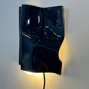 Puede incluir: Una l&aacute;mpara de pared con una pantalla de cer&aacute;mica azul marino oscuro brillante con una superficie irregular, plegada y ondulada. El accesorio emite un brillo c&aacute;lido y suave desde la parte inferior y tiene un cable de alimentaci&oacute;n negro.