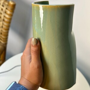 May include: A cylindrical ceramic vase with a smooth, glossy finish in a muted sage green color. The vase features a distinct vertical cutout along the top rim, creating an open, curved edge. The surface shows subtle tonal variations in the glaze.
