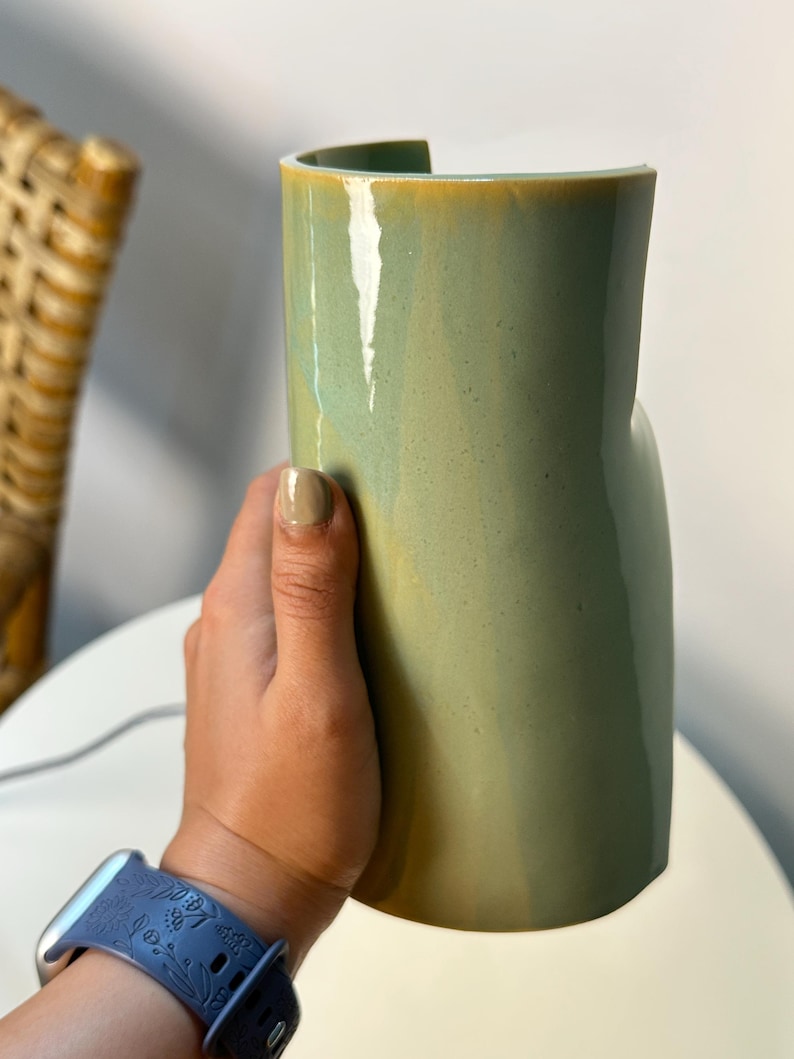 May include: A cylindrical ceramic vase with a smooth, glossy finish in a muted sage green color. The vase features a distinct vertical cutout along the top rim, creating an open, curved edge. The surface shows subtle tonal variations in the glaze.