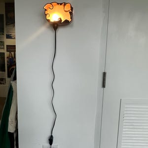 May include: A decorative wall sconce with a unique, organic shape. The sconce features a warm, orange glow emanating from within a dark brown frame. A black cord with a plug extends downward, connecting to a wall outlet.