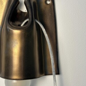 May include: A wall-mounted sconce featuring a dark bronze, metallic finish with a curved, folded design. The fixture includes a clear, transparent power cord threaded through the top and a single, round, frosted glass light bulb at the base.