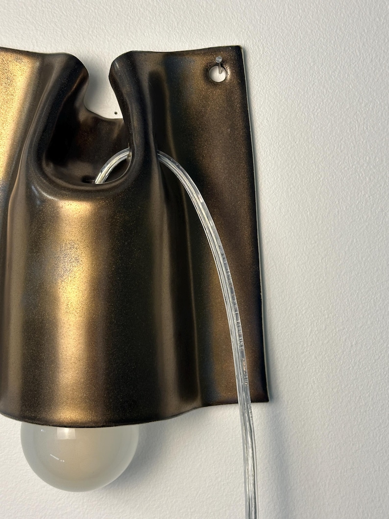 May include: A wall-mounted sconce featuring a dark bronze, metallic finish with a curved, folded design. The fixture includes a clear, transparent power cord threaded through the top and a single, round, frosted glass light bulb at the base.