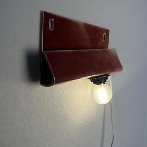 Może przedstawiać: Lampa ścienna z błyszczącą, ciemnoczerwoną ceramiczną oprawą o składanym, prostokątnym kształcie. Element posiada dwa pionowe otwory montażowe u g&oacute;ry oraz pojedynczą, okrągłą żar&oacute;wkę z matowego białego szkła umieszczoną u dołu konstrukcji.