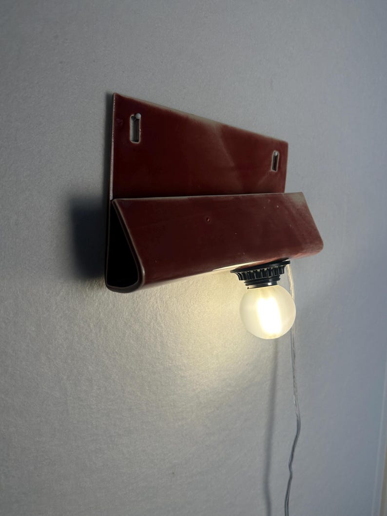 Może przedstawiać: Lampa ścienna z błyszczącą, ciemnoczerwoną ceramiczną oprawą o składanym, prostokątnym kształcie. Element posiada dwa pionowe otwory montażowe u g&oacute;ry oraz pojedynczą, okrągłą żar&oacute;wkę z matowego białego szkła umieszczoną u dołu konstrukcji.