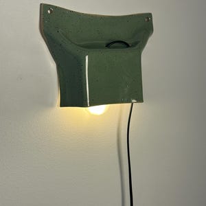 May include: A wall-mounted ceramic lamp with a glossy sage green finish and dark speckles. The fixture features a curved, flared top section with two mounting holes and a downward-facing opening that emits a warm light from a single bulb.