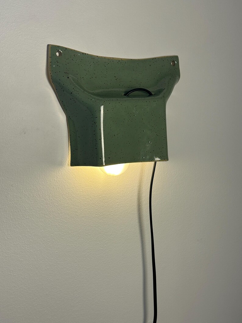 May include: A wall-mounted ceramic lamp with a glossy sage green finish and dark speckles. The fixture features a curved, flared top section with two mounting holes and a downward-facing opening that emits a warm light from a single bulb.