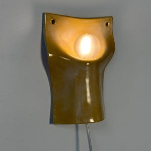 May include: A mustard yellow ceramic wall sconce with a curved, concave silhouette. The fixture features a central circular opening holding a glowing, frosted glass globe bulb. Two small mounting holes are visible at the top corners of the piece.