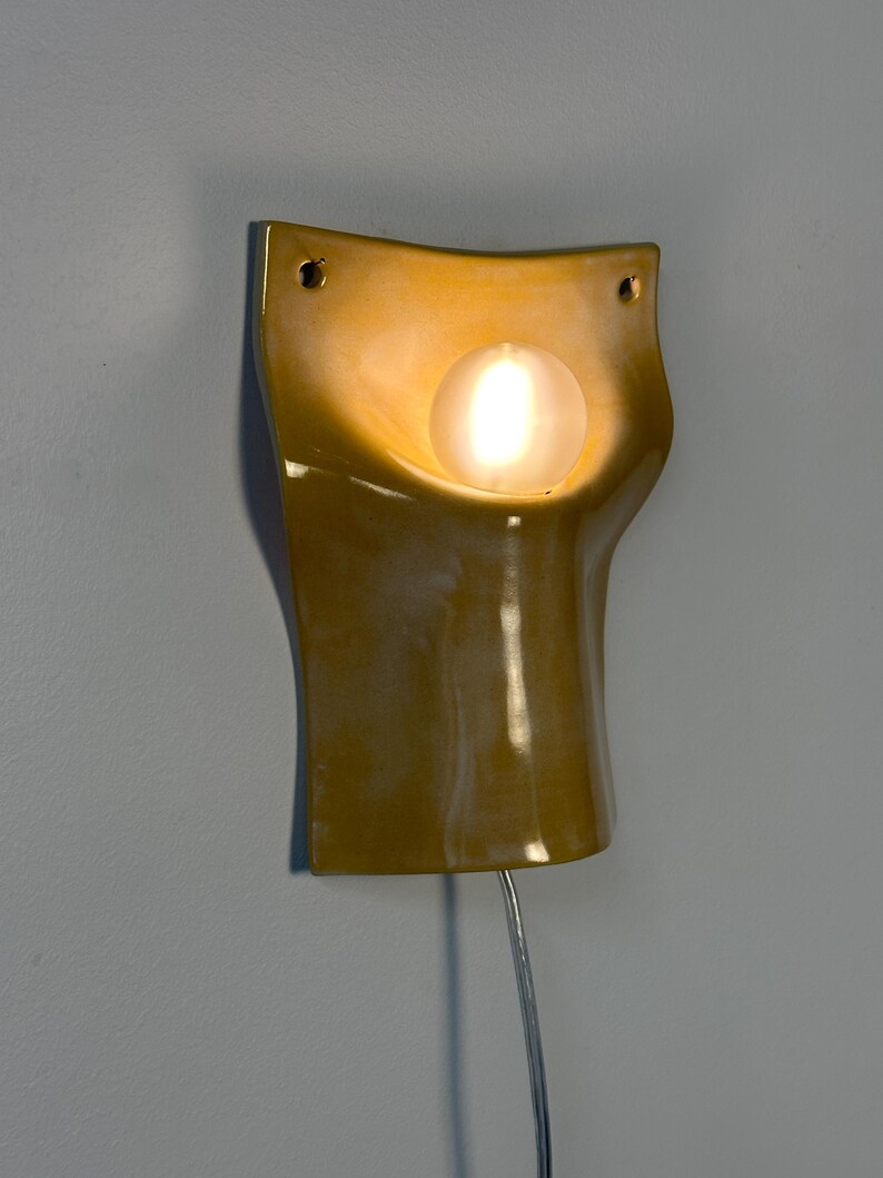 May include: A mustard yellow ceramic wall sconce with a curved, concave silhouette. The fixture features a central circular opening holding a glowing, frosted glass globe bulb. Two small mounting holes are visible at the top corners of the piece.