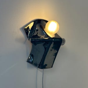 May include: A wall-mounted lamp featuring a glossy, dark charcoal-colored ceramic base with an irregular, folded shape. A single, round, frosted glass light bulb is attached to the upper section, emitting a warm, soft glow against the wall.