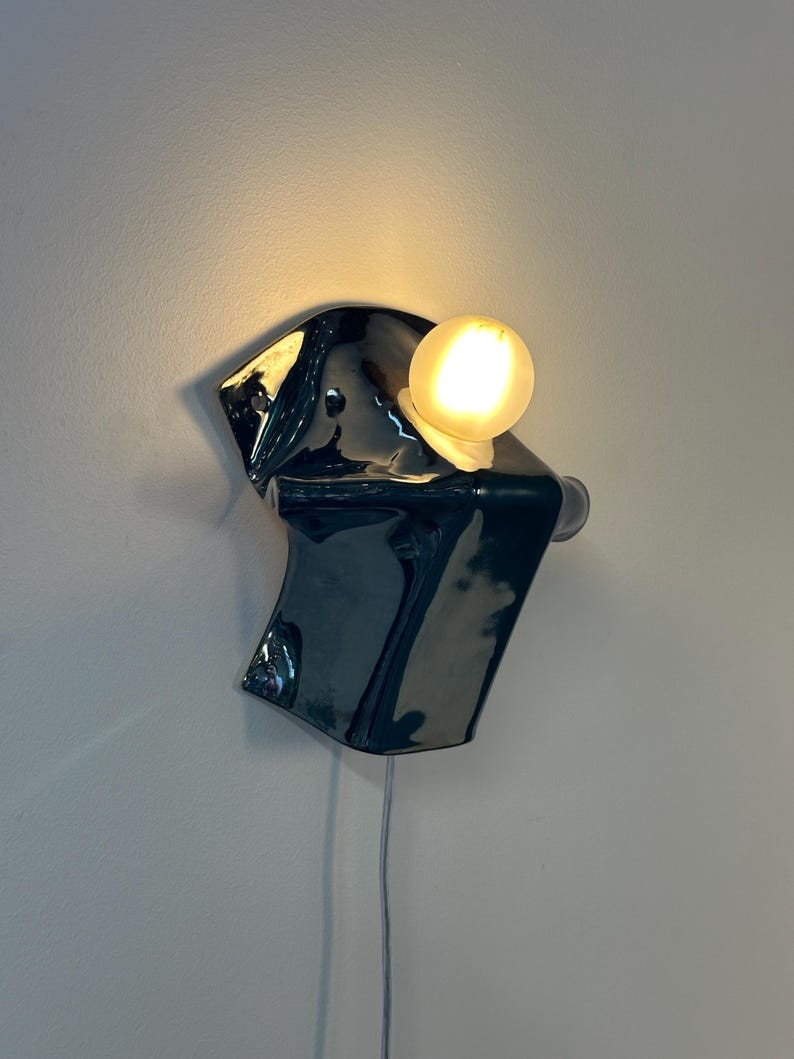 May include: A wall-mounted lamp featuring a glossy, dark charcoal-colored ceramic base with an irregular, folded shape. A single, round, frosted glass light bulb is attached to the upper section, emitting a warm, soft glow against the wall.