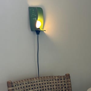 Pu&ograve; includere: Una lampada da parete con un paralume in ceramica verde lucido e curvo che racchiude parzialmente una lampadina accesa. L'apparecchio include un portalampada nero e un sottile cavo di alimentazione nero che pende verticalmente lungo la parete.