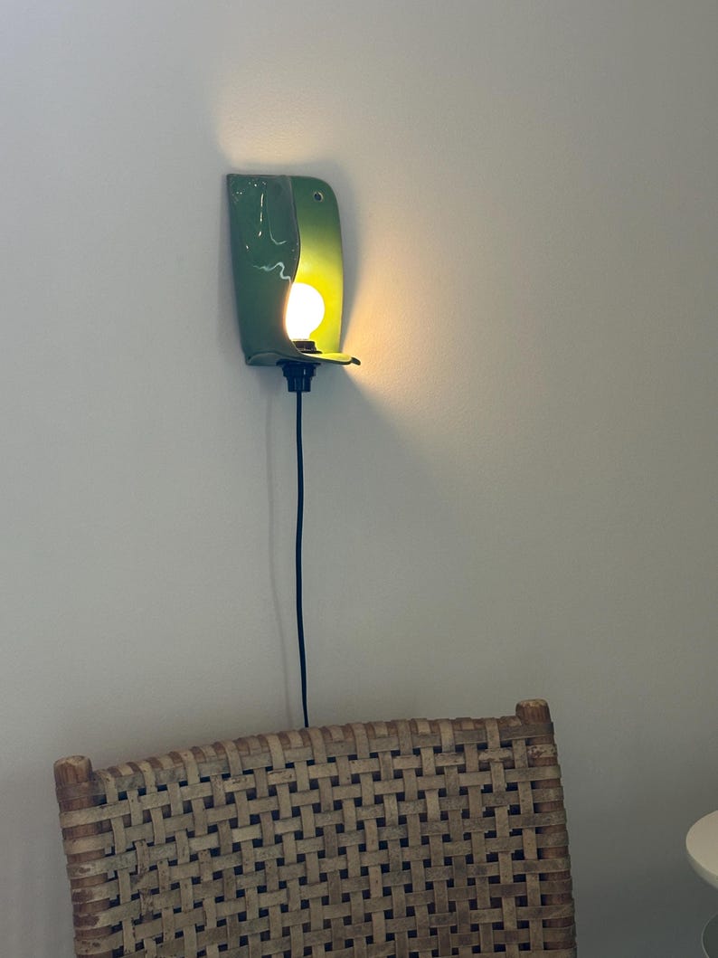 Pu&ograve; includere: Una lampada da parete con un paralume in ceramica verde lucido e curvo che racchiude parzialmente una lampadina accesa. L'apparecchio include un portalampada nero e un sottile cavo di alimentazione nero che pende verticalmente lungo la parete.