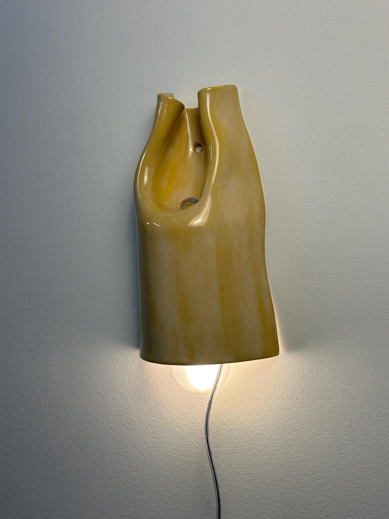 Pu&ograve; includere: Una lampada da parete in ceramica con una finitura lucida color giallo senape. L'oggetto presenta una forma organica, piegata e asimmetrica con un centro cavo. Una singola lampadina rotonda &egrave; visibile in basso, emettendo una luce calda.