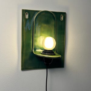 May include: A wall-mounted ceramic light fixture featuring a glossy, deep green square backplate with a central arched indentation. A small, round, frosted glass bulb sits on a black socket base extending from the center of the curved shelf.