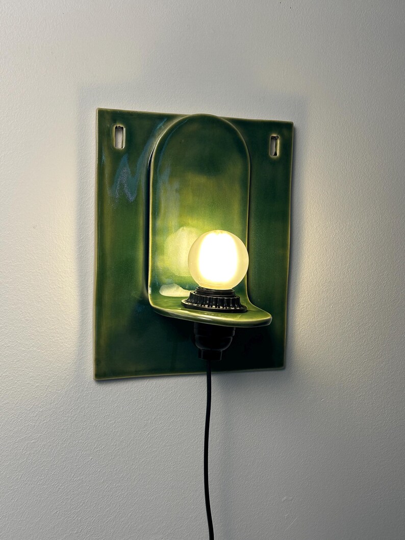 May include: A wall-mounted ceramic light fixture featuring a glossy, deep green square backplate with a central arched indentation. A small, round, frosted glass bulb sits on a black socket base extending from the center of the curved shelf.