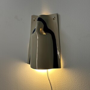 May include: A wall-mounted ceramic sconce featuring a glossy off-white finish with a dark blue vertical stripe. The fixture has a curved, folded design that emits a warm, glowing light from the top and bottom, and includes a clear power cord.