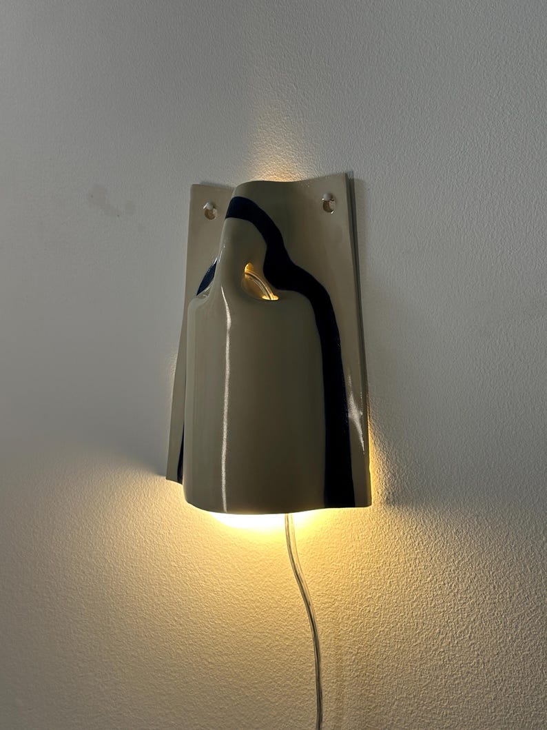 May include: A wall-mounted ceramic sconce featuring a glossy off-white finish with a dark blue vertical stripe. The fixture has a curved, folded design that emits a warm, glowing light from the top and bottom, and includes a clear power cord.