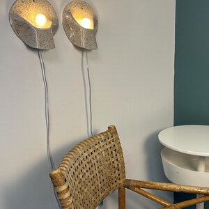 May include: Two wall-mounted sconces with a speckled off-white finish and a curved, shell-like shape. Each fixture contains a single glowing light bulb and features a thin, light-coloured power cord extending downwards from the base of the lamp.