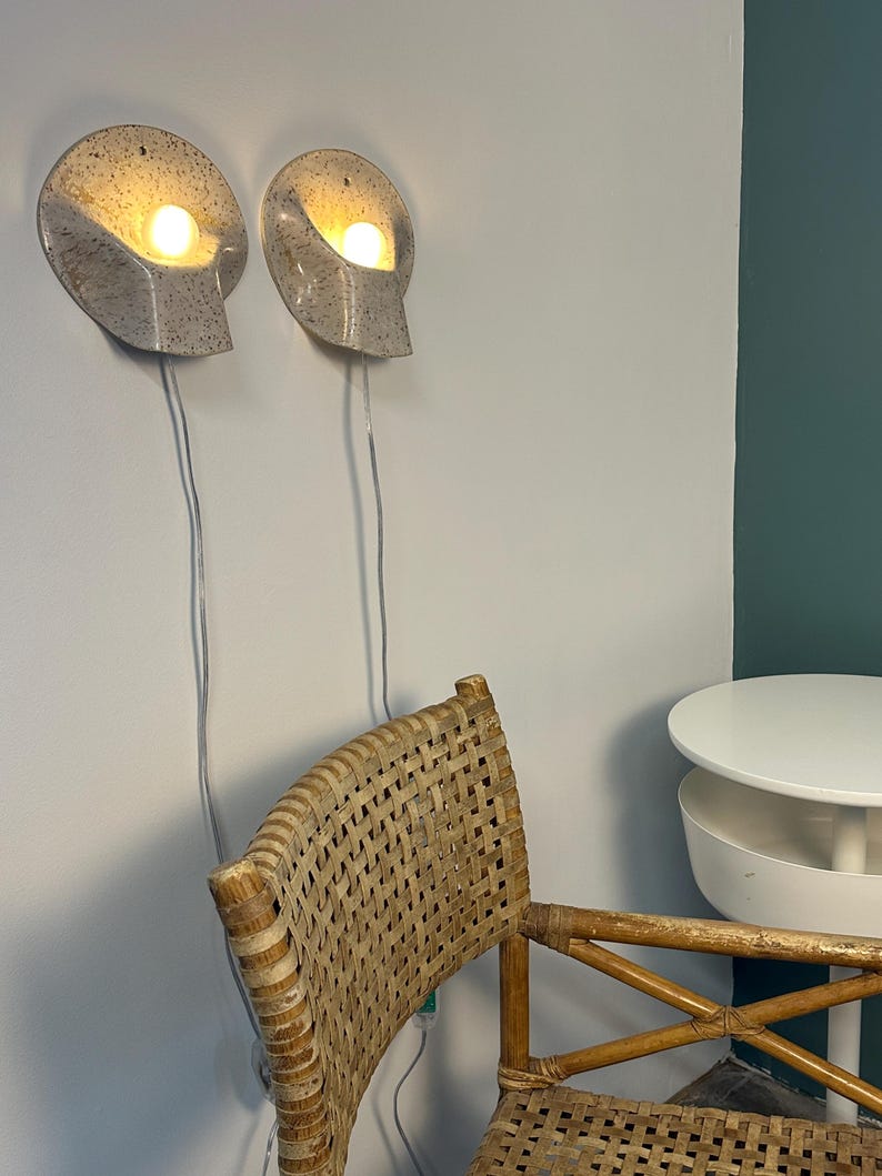 May include: Two wall-mounted sconces with a speckled off-white finish and a curved, shell-like shape. Each fixture contains a single glowing light bulb and features a thin, light-coloured power cord extending downwards from the base of the lamp.