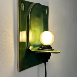 May include: A wall-mounted ceramic sconce featuring a glossy, deep green glaze. The fixture has a rectangular backplate with an arched center and a small, horizontal shelf supporting a round, glowing light bulb in a black socket with a black cord.