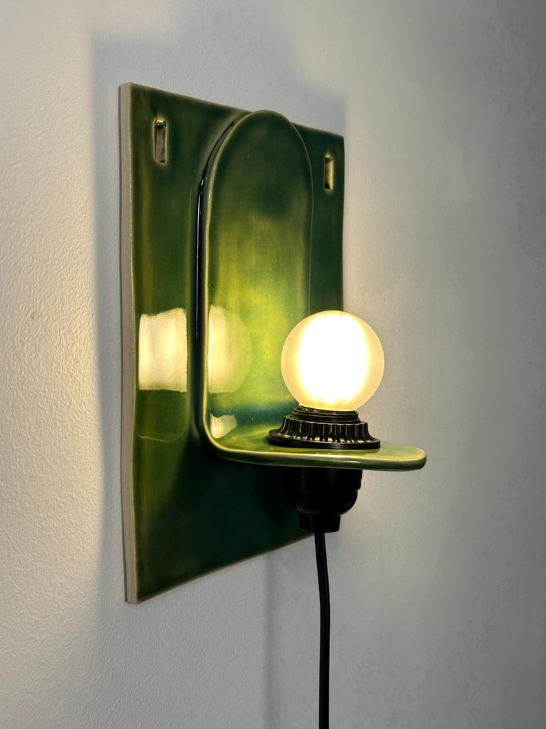 May include: A wall-mounted ceramic sconce featuring a glossy, deep green glaze. The fixture has a rectangular backplate with an arched center and a small, horizontal shelf supporting a round, glowing light bulb in a black socket with a black cord.