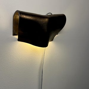 May include: A wall-mounted lamp featuring a dark, curved exterior shell with a metallic gold interior finish. The fixture emits a warm, downward-facing glow and includes a thin, light-coloured power cord extending from the bottom of the unit.