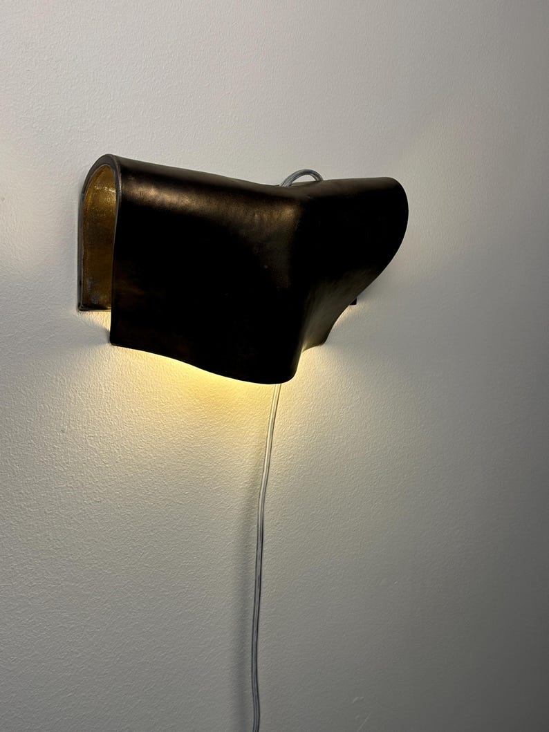 May include: A wall-mounted lamp featuring a dark, curved exterior shell with a metallic gold interior finish. The fixture emits a warm, downward-facing glow and includes a thin, light-coloured power cord extending from the bottom of the unit.