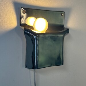 Peut inclure: Une applique murale en c&eacute;ramique avec une finition vert fonc&eacute; brillante. Le luminaire pr&eacute;sente un corps rectangulaire incurv&eacute; avec une section sup&eacute;rieure en retrait contenant une ampoule sph&eacute;rique lumineuse. Un fin cordon blanc d&eacute;passe du bas.