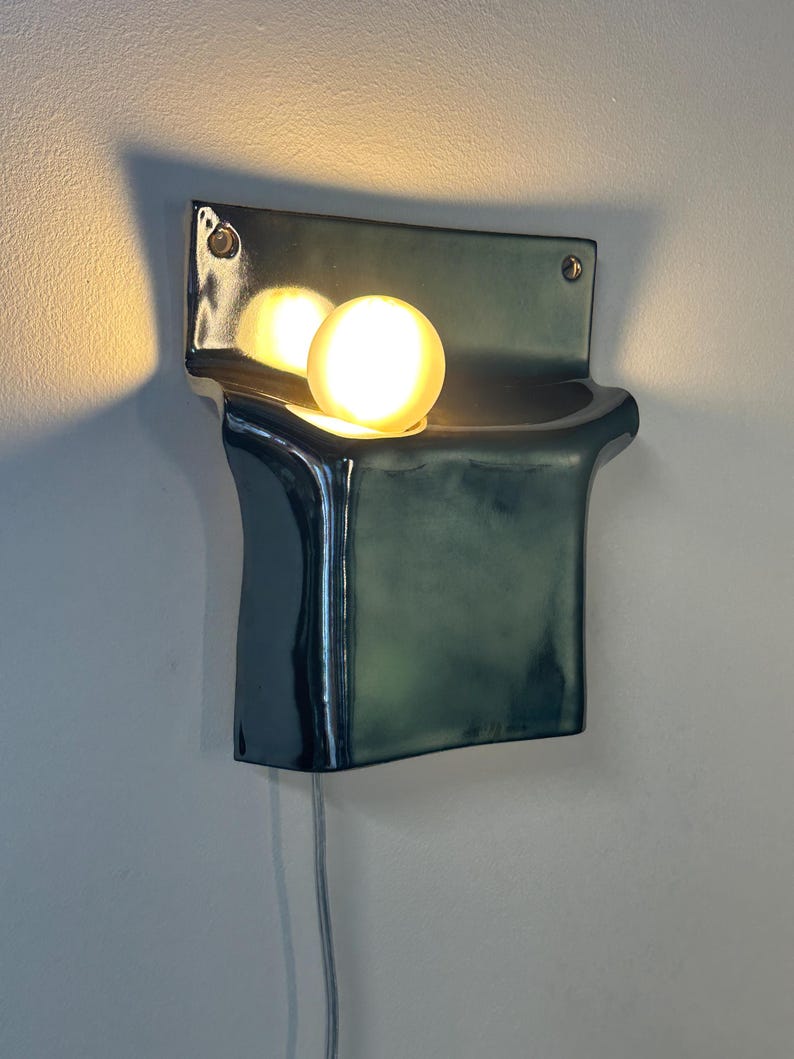 Peut inclure: Une applique murale en c&eacute;ramique avec une finition vert fonc&eacute; brillante. Le luminaire pr&eacute;sente un corps rectangulaire incurv&eacute; avec une section sup&eacute;rieure en retrait contenant une ampoule sph&eacute;rique lumineuse. Un fin cordon blanc d&eacute;passe du bas.