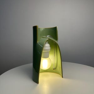 May include: A sage green ceramic table lamp with a curved, architectural structure. The lamp features a glossy finish and an open, arched design that houses a single white light bulb socket and a glowing, frosted light bulb suspended inside.