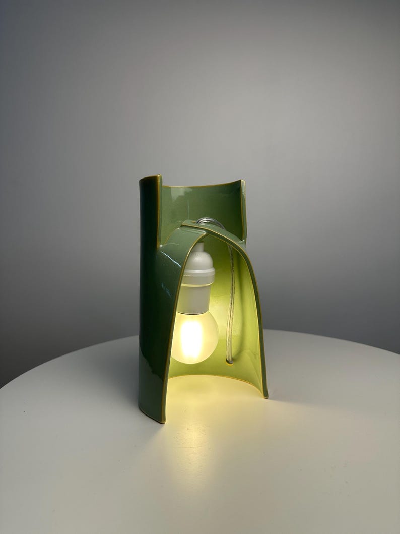 May include: A sage green ceramic table lamp with a curved, architectural structure. The lamp features a glossy finish and an open, arched design that houses a single white light bulb socket and a glowing, frosted light bulb suspended inside.