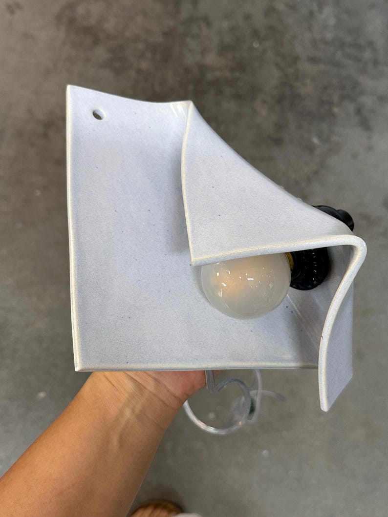 May include: A wall-mounted light fixture featuring a folded, light gray ceramic shade with a speckled texture. The fixture includes a single round light bulb set into a black socket, with a small circular hole at the top corner for mounting.