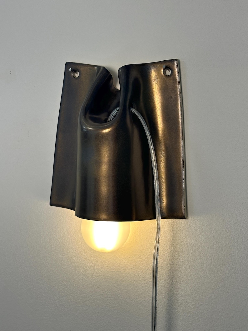May include: A wall-mounted lamp featuring a dark, glossy, curved ceramic shade with two mounting holes at the top corners. A single round, glowing light bulb is positioned at the bottom, and a clear electrical cord hangs down the right side.