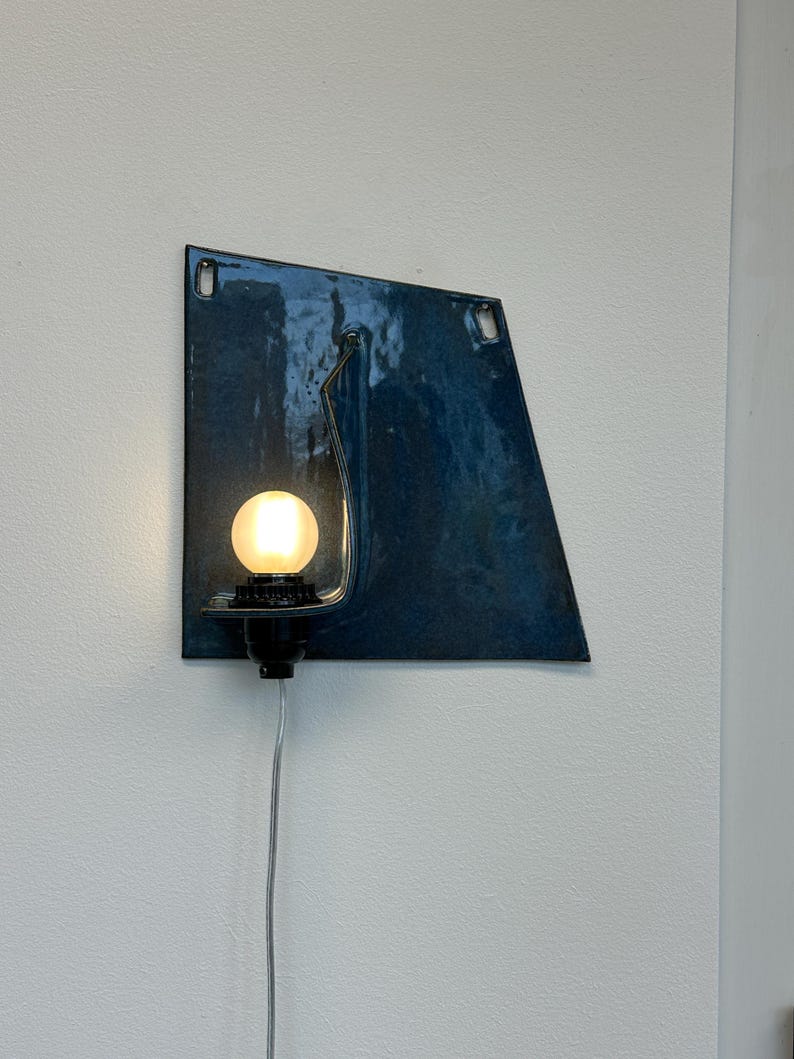 May include: A modern wall sconce with a dark blue, geometric metal shade. The sconce features a black bulb socket and a glowing, round light bulb. A silver cord extends from the base.