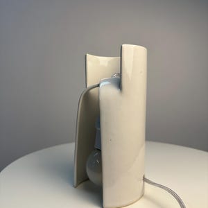 May include: A cream-colored ceramic table lamp with a curved, cylindrical structure that partially encloses a single light bulb. The lamp features a smooth, glossy finish and includes a thin, light-colored electrical cord extending from the base.