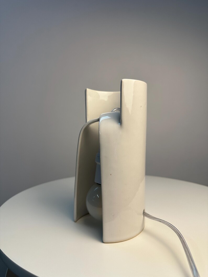 May include: A cream-colored ceramic table lamp with a curved, cylindrical structure that partially encloses a single light bulb. The lamp features a smooth, glossy finish and includes a thin, light-colored electrical cord extending from the base.