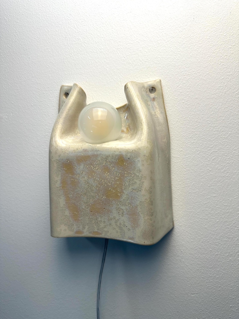 May include: A wall-mounted ceramic light fitting with a glossy, off-white glaze and mottled tan texture. The rectangular body features two top openings, one holding a round, frosted glass light bulb. A thin electrical cord extends from the bottom.