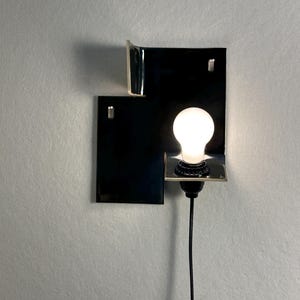 Pode incluir: Uma arandela de parede moderna com um design retangular preto e uma l&acirc;mpada brilhante. A arandela tem uma est&eacute;tica minimalista, com um cabo preto e uma forma geom&eacute;trica simples. A l&acirc;mpada &eacute; redonda e emite um brilho quente.