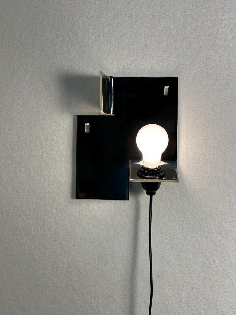 Pode incluir: Uma arandela de parede moderna com um design retangular preto e uma l&acirc;mpada brilhante. A arandela tem uma est&eacute;tica minimalista, com um cabo preto e uma forma geom&eacute;trica simples. A l&acirc;mpada &eacute; redonda e emite um brilho quente.